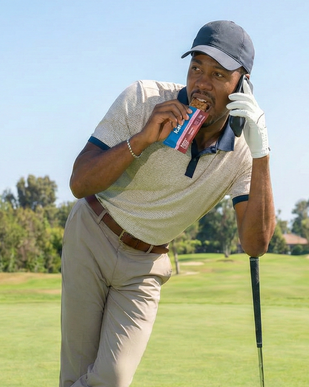 Man on a golf course eating a Kailo protein bar as a snack and talking on a phone