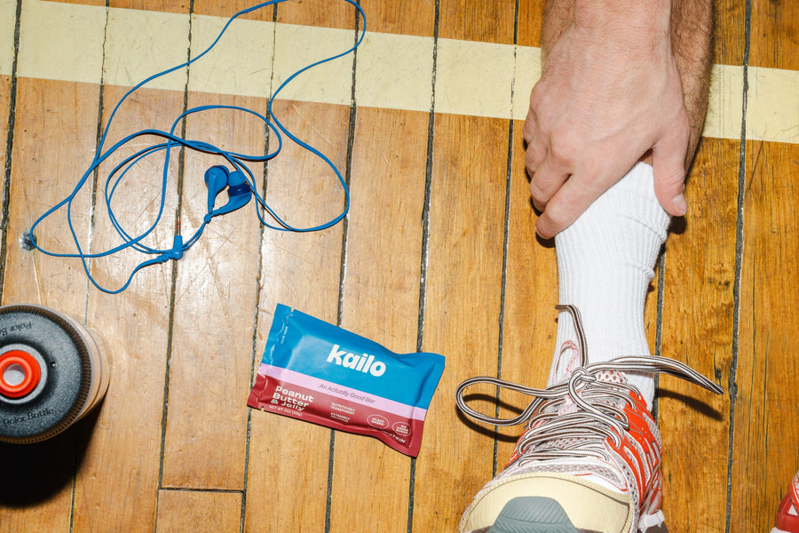 A Kailo Peanut Butter and Jelly bar on a gym floor beside a water bottle and earbuds while an athlete laces up